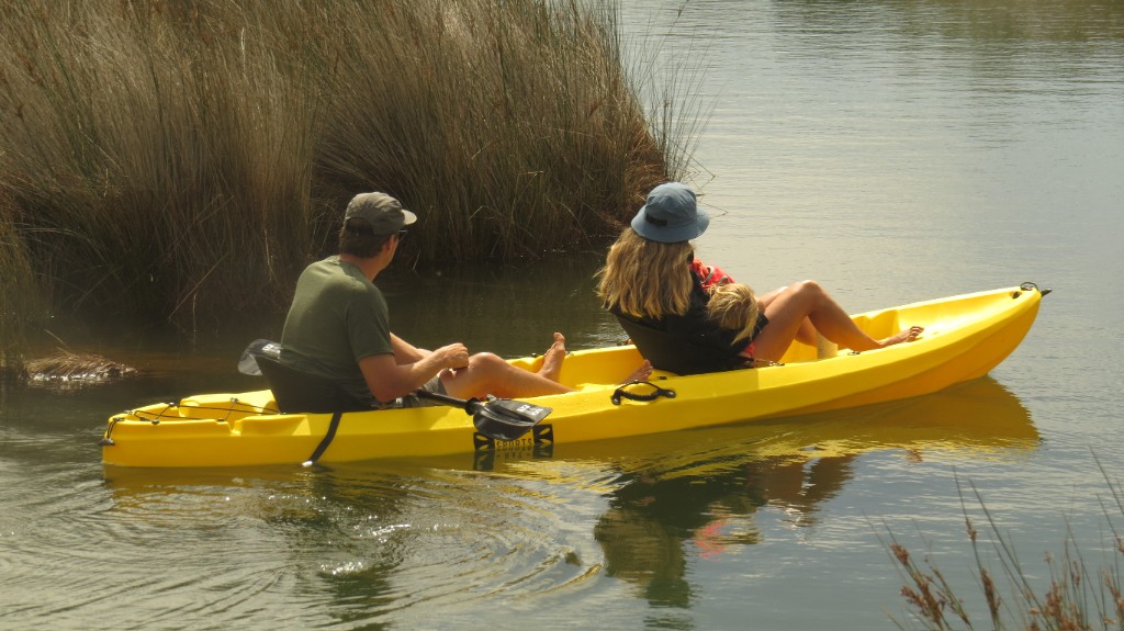 Family of three in a bright yellow tandem kayak on calm water near tall marsh grasses