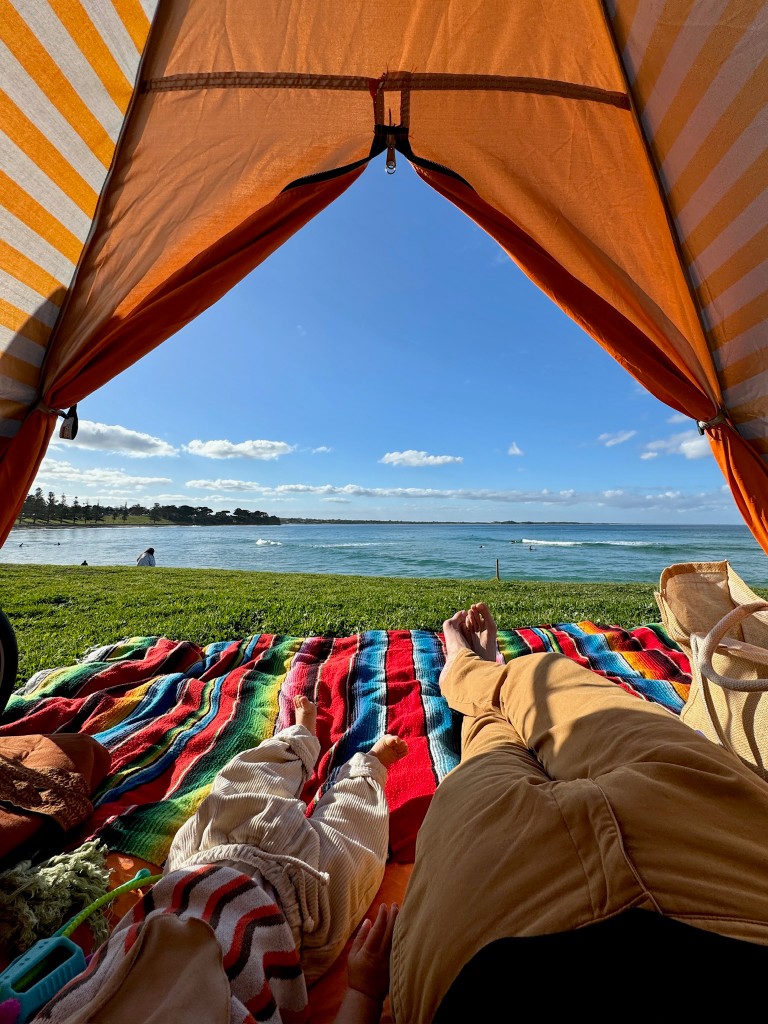 View from inside a striped tent toward the ocean, with a parent and child resting on a colourful blanket